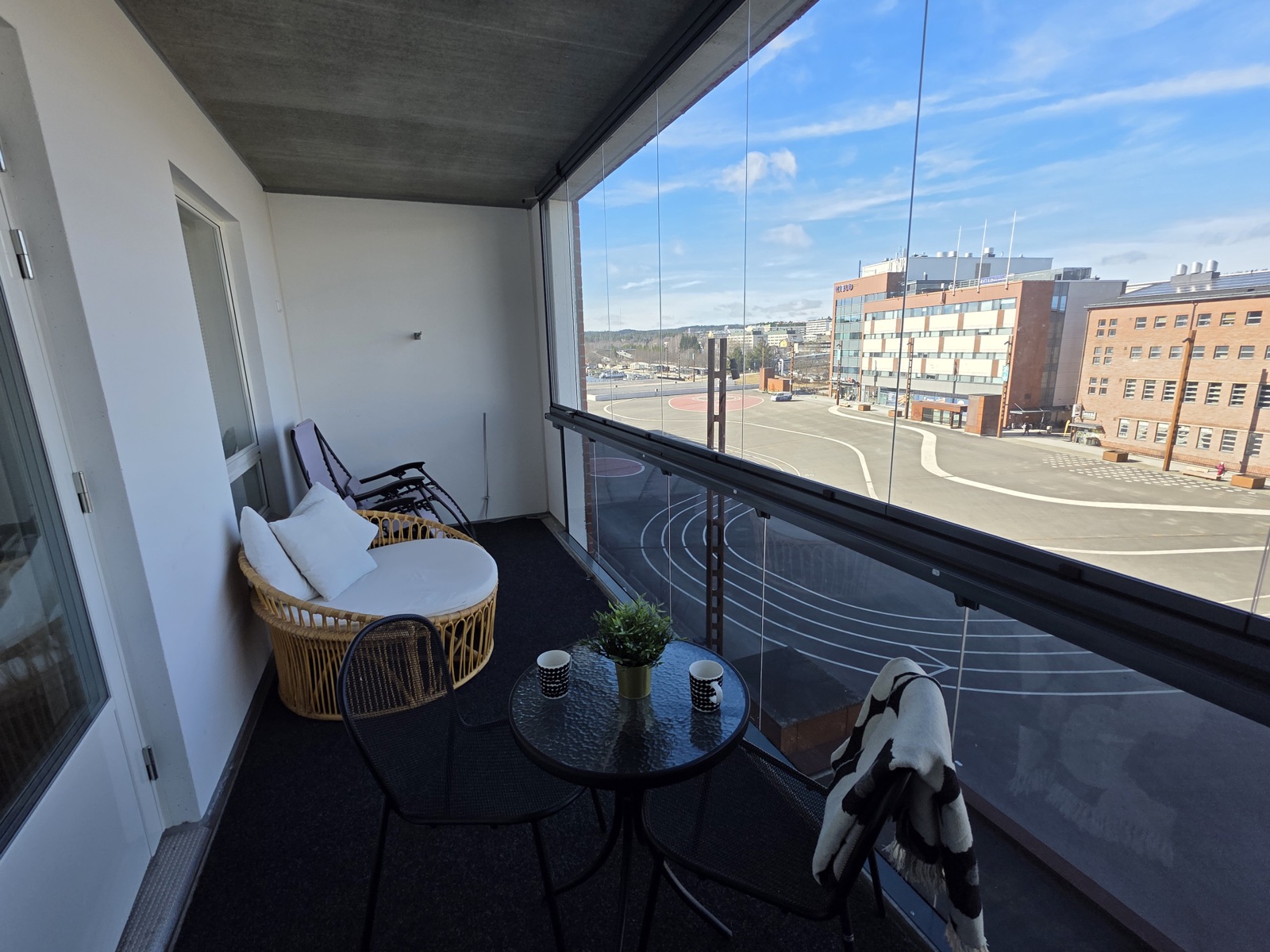 Cozy balcony seating area with a wicker chair, black metal chairs, and a round glass table, overlooking a city street and buildings beyond.